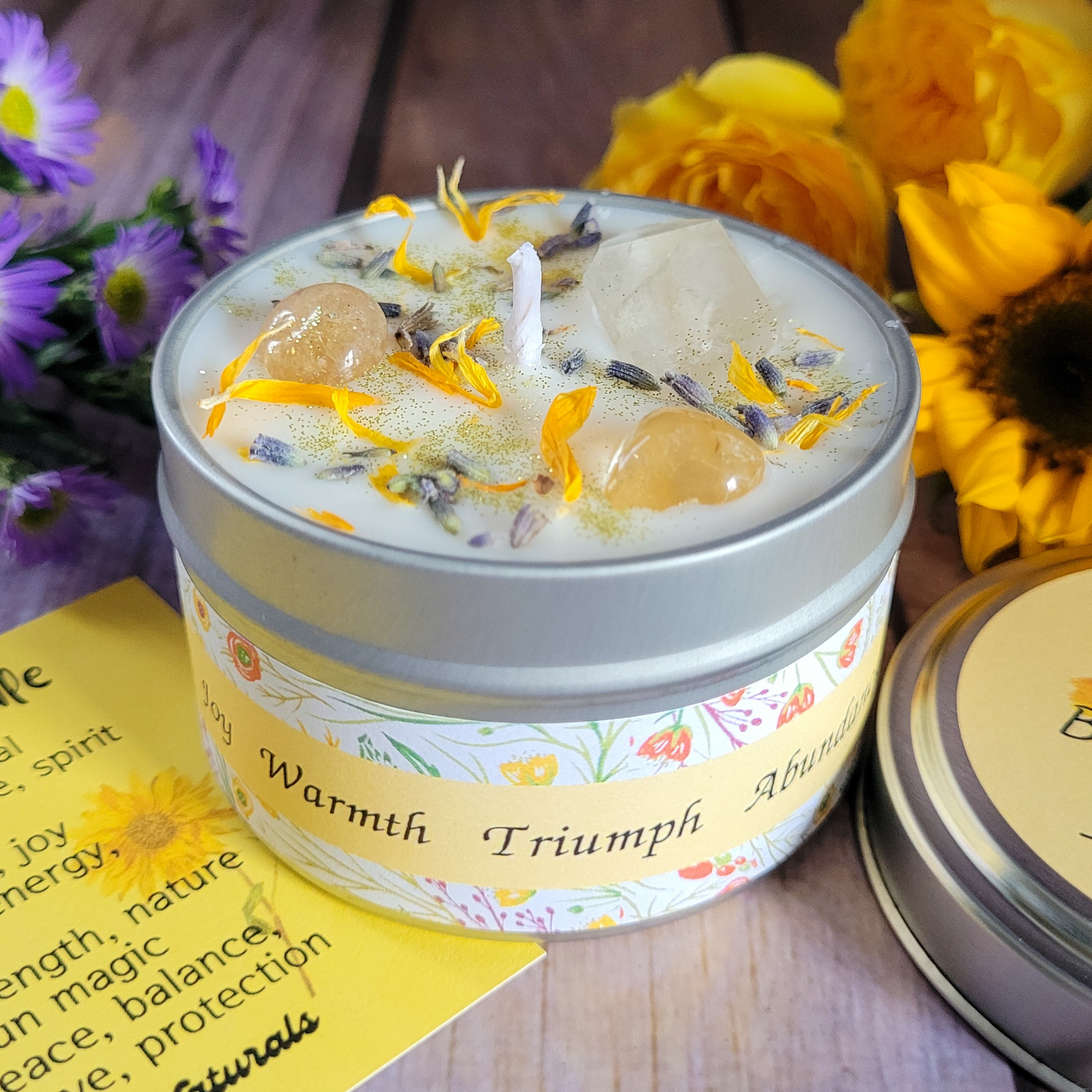 Candle in a tin with floral elements and a label, surrounded by flowers on a wooden surface.
