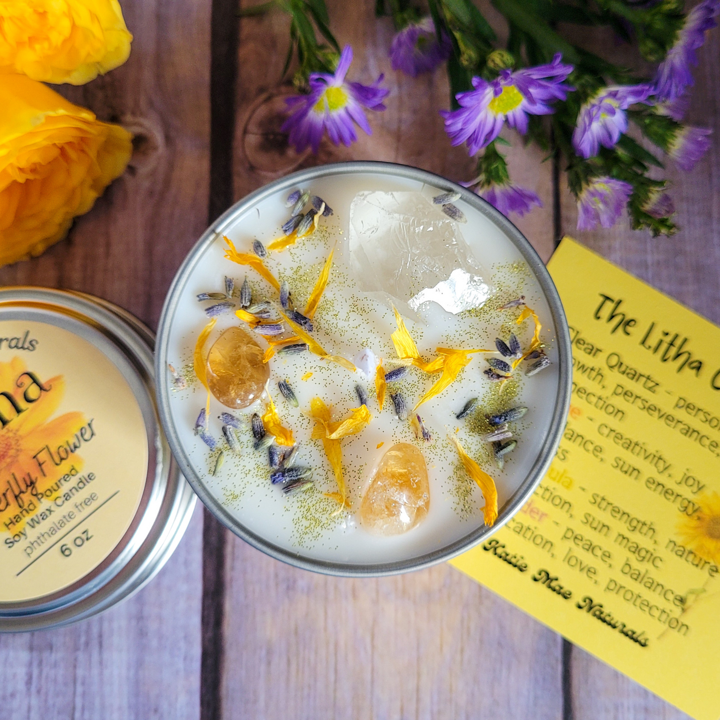 Candle in a tin with floral elements on a wooden surface with flowers and a label in the background.