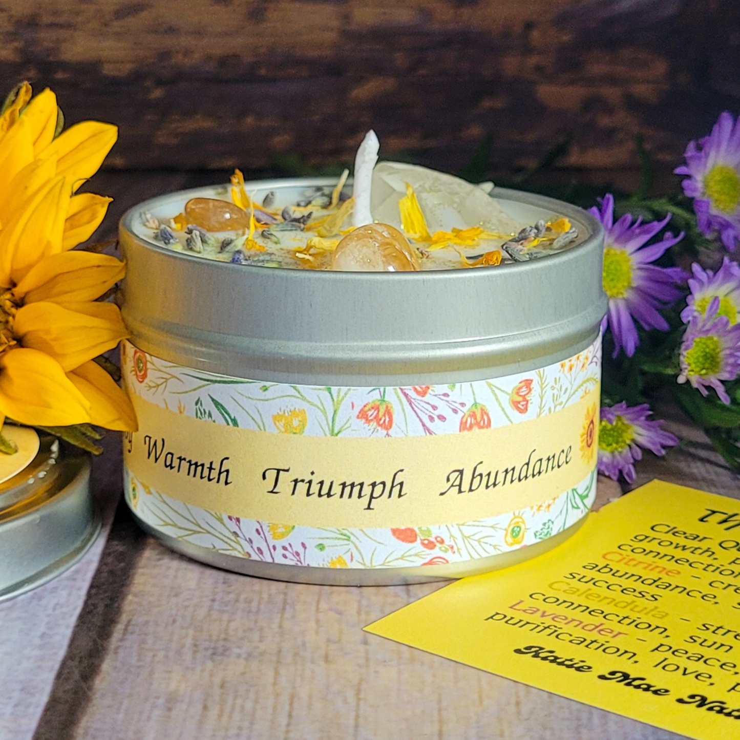 Candle in a tin with decorative label surrounded by flowers on a wooden surface