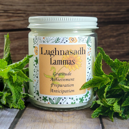 Candle jar with 'Lughnasadh Lammas' label surrounded by green leaves on a wooden surface