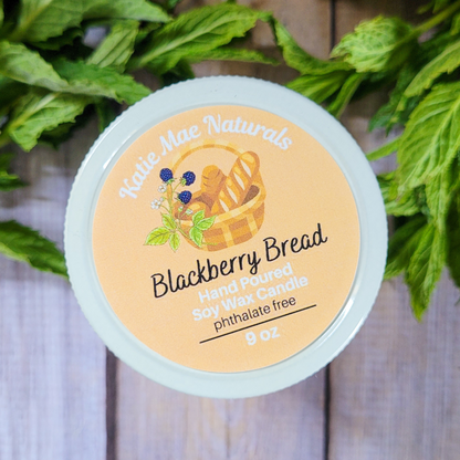 Hand poured soy wax candle labeled 'Blackberry Bread' by Kettle Mae Naturals on a wooden background with greenery.