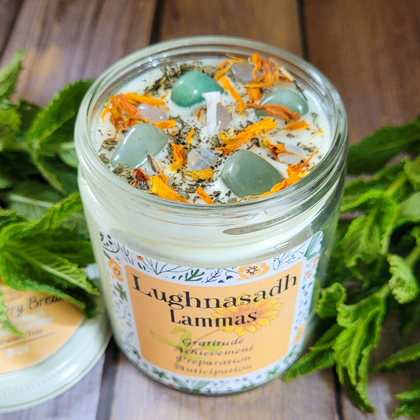 Candle jar with decorative elements and 'Lughnasadh Lammas' label on a wooden surface with greenery.