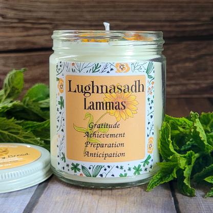 Candle in a jar with 'Lughnasadh Lammas' label surrounded by greenery on a wooden surface