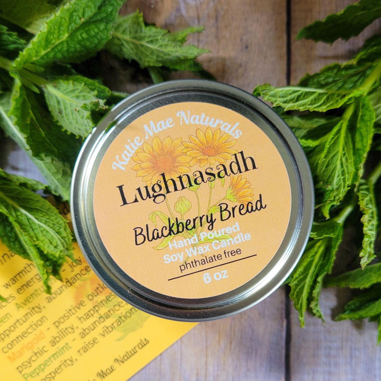 Candle jar labeled 'Lughnasadh Blackberry Bread' surrounded by green leaves on a wooden surface.