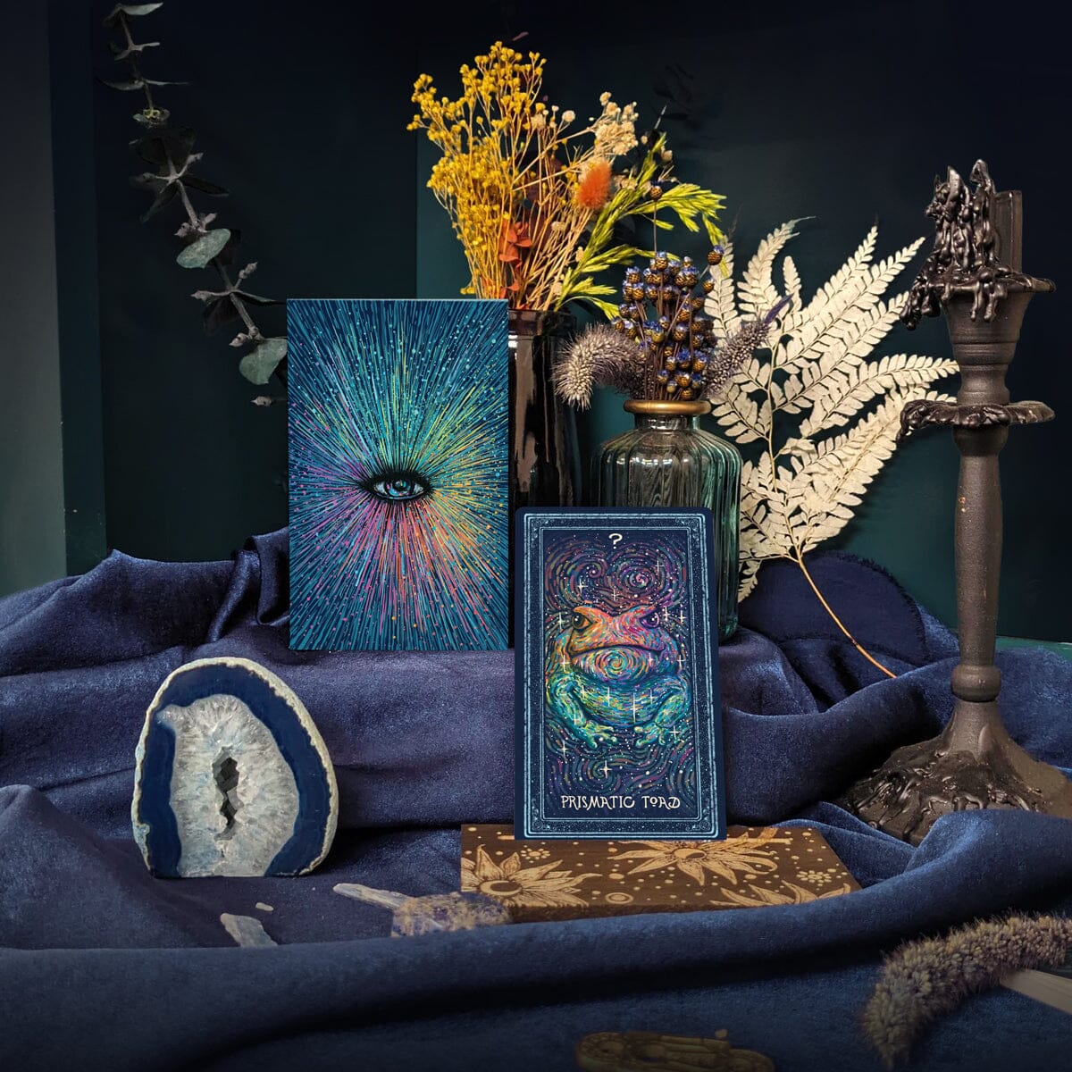 A styled product display featuring a tarot card deck box labeled 'MEMENTO TYRANNIS' and a matching illustrated card, surrounded by dried flowers, a glass vase, a candle holder, a stone slice, and dark blue fabric against a dark teal background.
