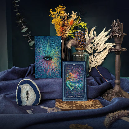 A styled product display featuring a tarot card deck box labeled 'MEMENTO TYRANNIS' and a matching illustrated card, surrounded by dried flowers, a glass vase, a candle holder, a stone slice, and dark blue fabric against a dark teal background.