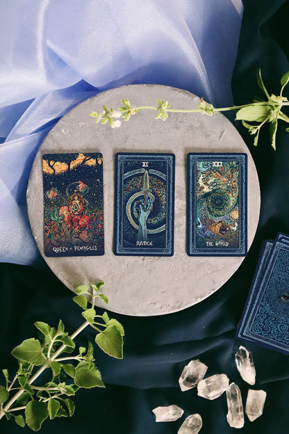 Three tarot cards on a round stone surface with crystals and greenery, against a dark fabric background.