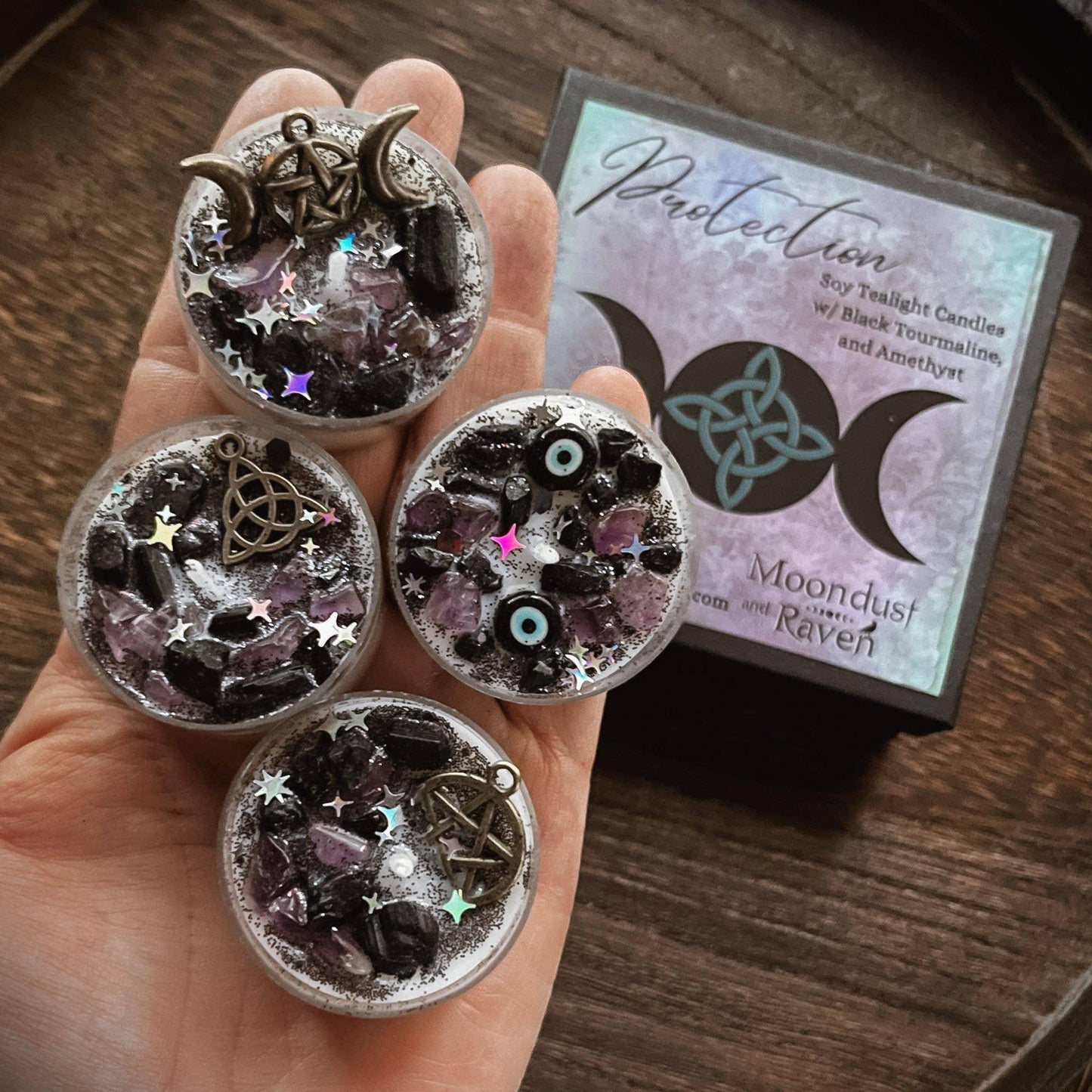 Four small round silver wax melts tins filled with black and purple crystals, stars, and moon symbols, held in a hand next to their branded packaging box on a wooden surface.