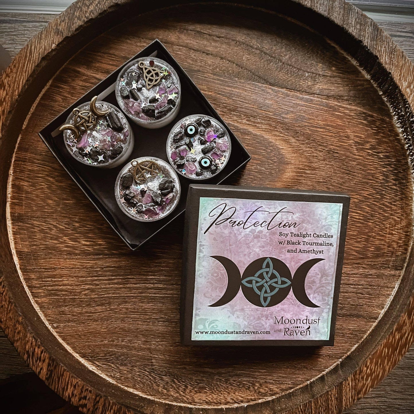 A boxed set of four moon phase soy candles in a black tray, shown next to the product card on a round wooden tray. The card reads 'Protection Soy Twilight Candles w/ black Traumatization and Ambition' and shows the brand name 'Moondust Rituals'.
