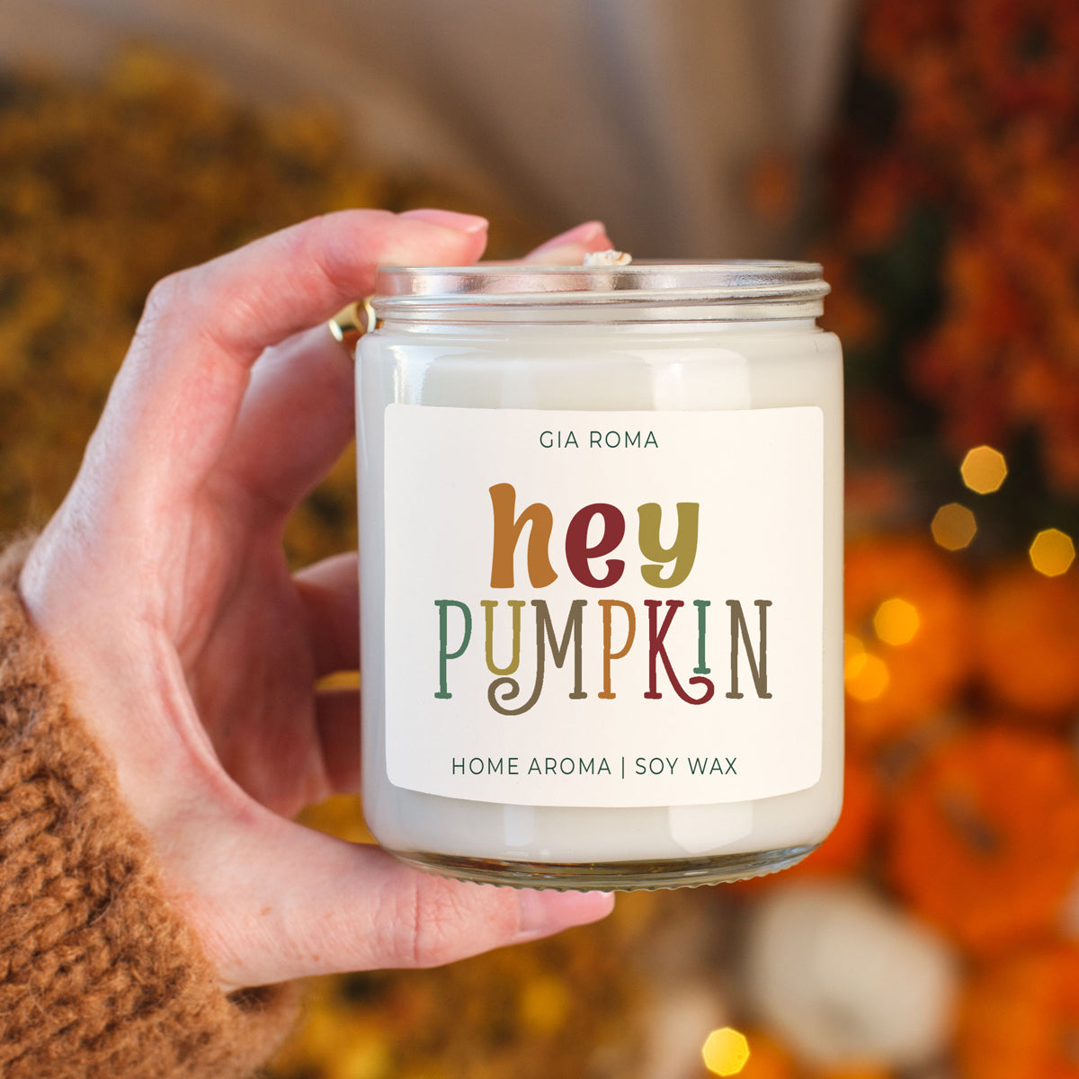 Hand holding a candle labeled 'hey pumpkin' with a blurred autumn background