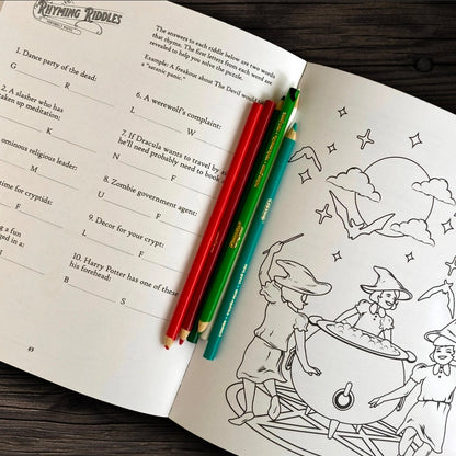An open black-and-white coloring book titled 'Cryptic Riddles' on a dark wooden table, with three colored pencils placed across the pages.