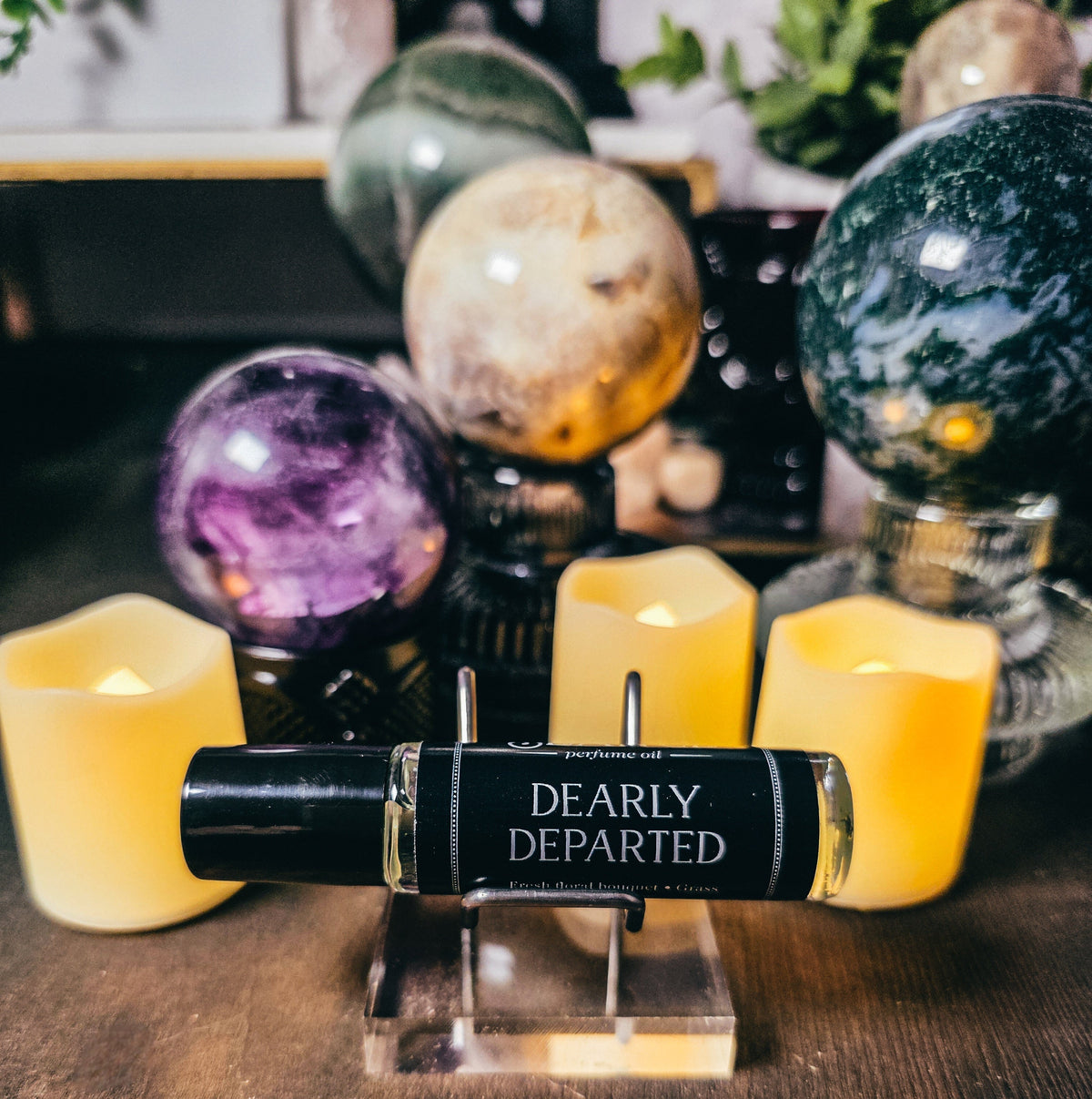Roll-on bottle labeled 'Dearly Departed' on a stand with candles and crystal balls in the background.