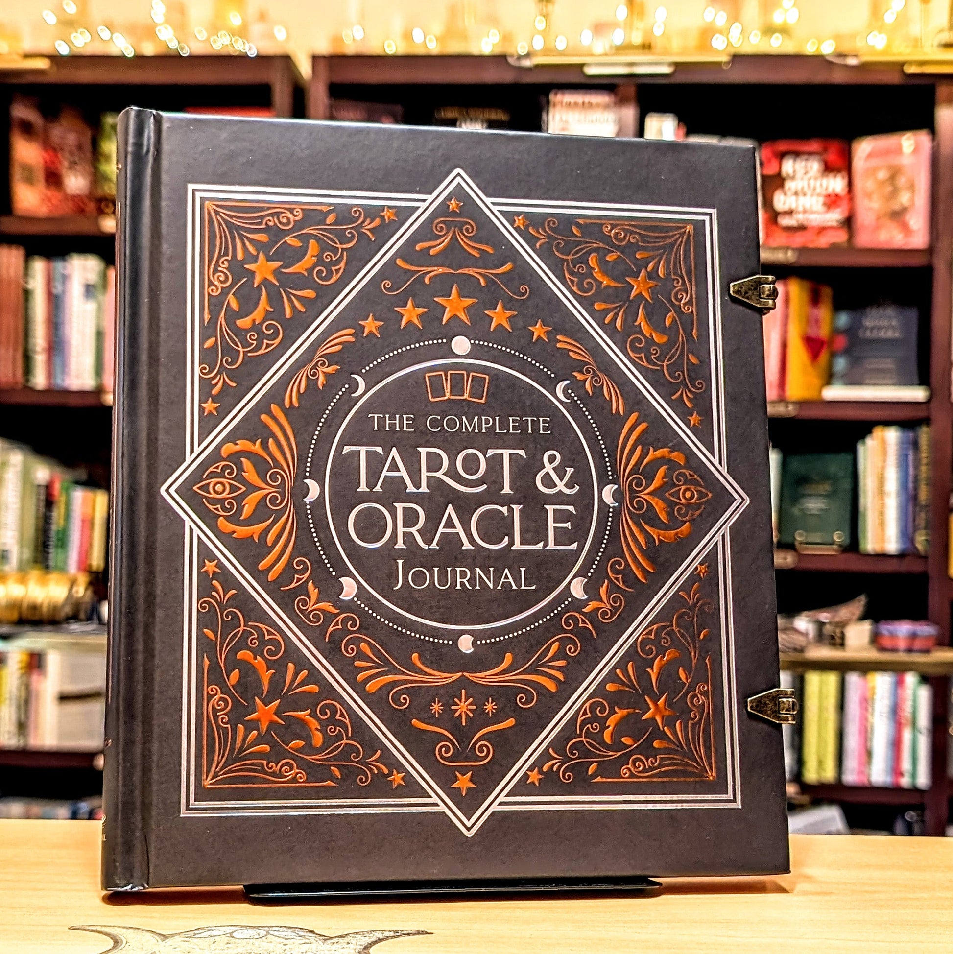 Decorative book titled 'The Complete Tarot & Oracle Journal' on a wooden surface with a bookshelf background.