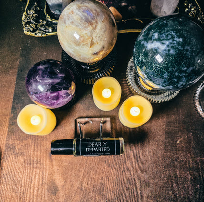 A roll-on perfume bottle labeled 'The Dearly Departed' lies on a wooden surface surrounded by crystals, candles, and decorative objects. The bottle has a black cap and body with white text, and the scene is styled in a mystical, altar-like arrangement.