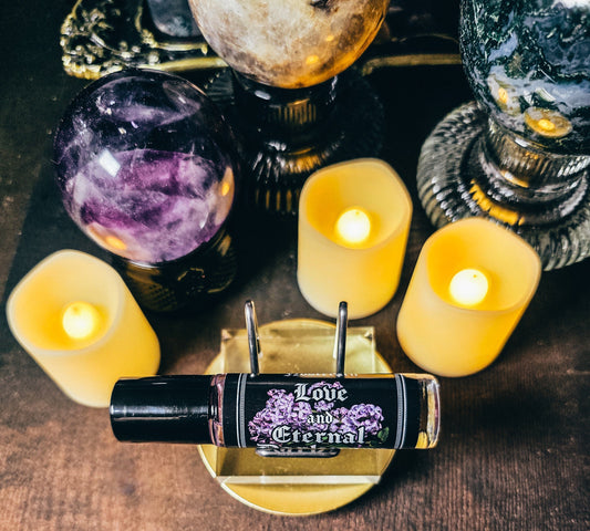 A small black-and-clear roller bottle labeled 'Love Eternal' sits on a gold stand on a wooden surface, surrounded by glowing yellow candles, crystals, and decorative objects.