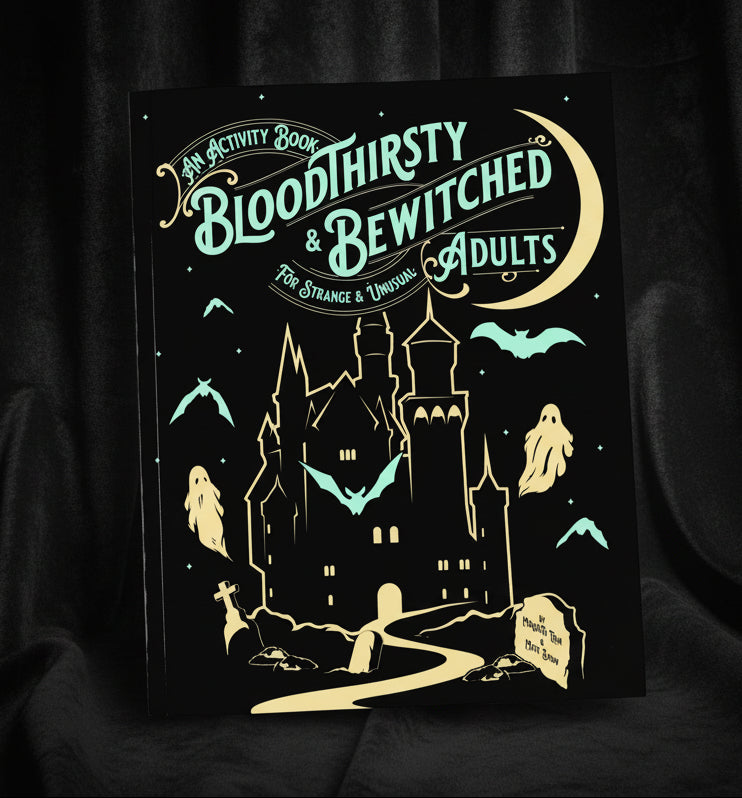 An activity book titled 'Bloodthirsty & Bewitched Adults' with a black cover, teal and gold illustration of a haunted house, bats, ghosts, and a crescent moon, styled on a mint background with ribbon and patterned paper props.