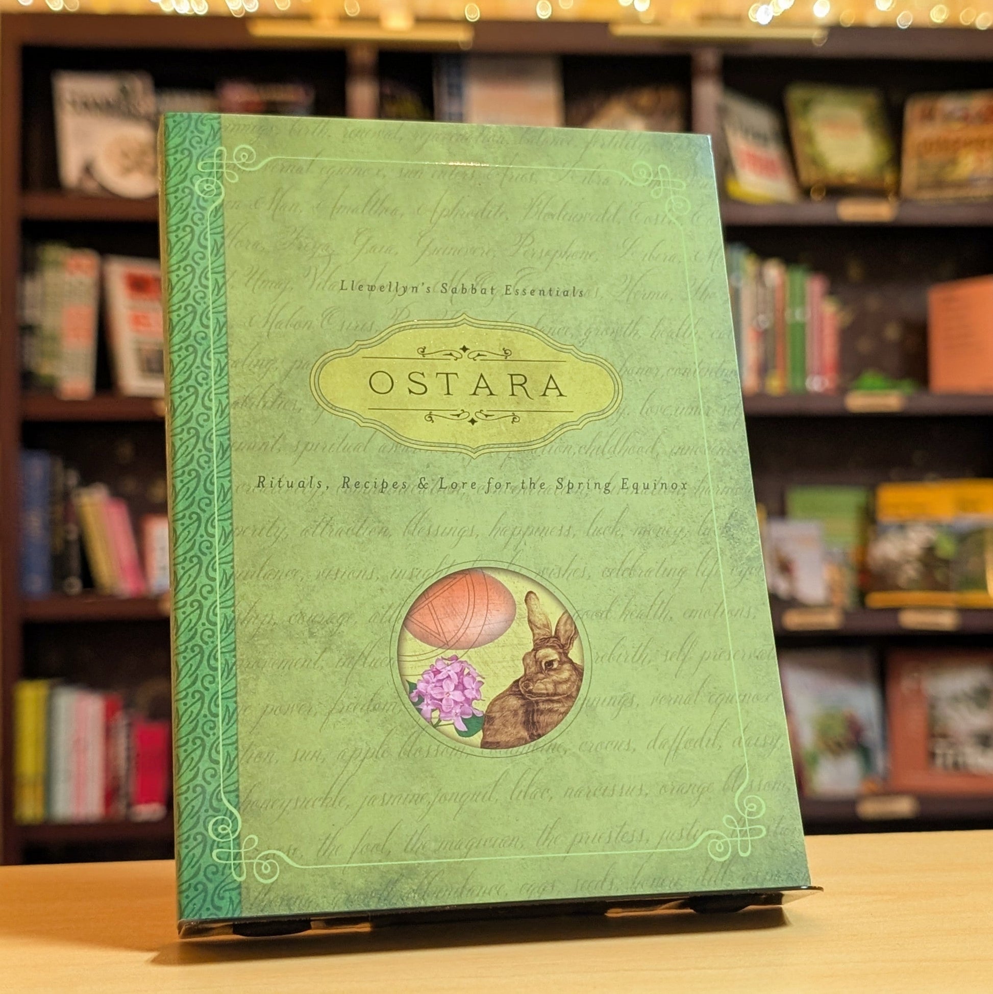 Green book titled 'Ostara' on a wooden surface with a bookshelf background