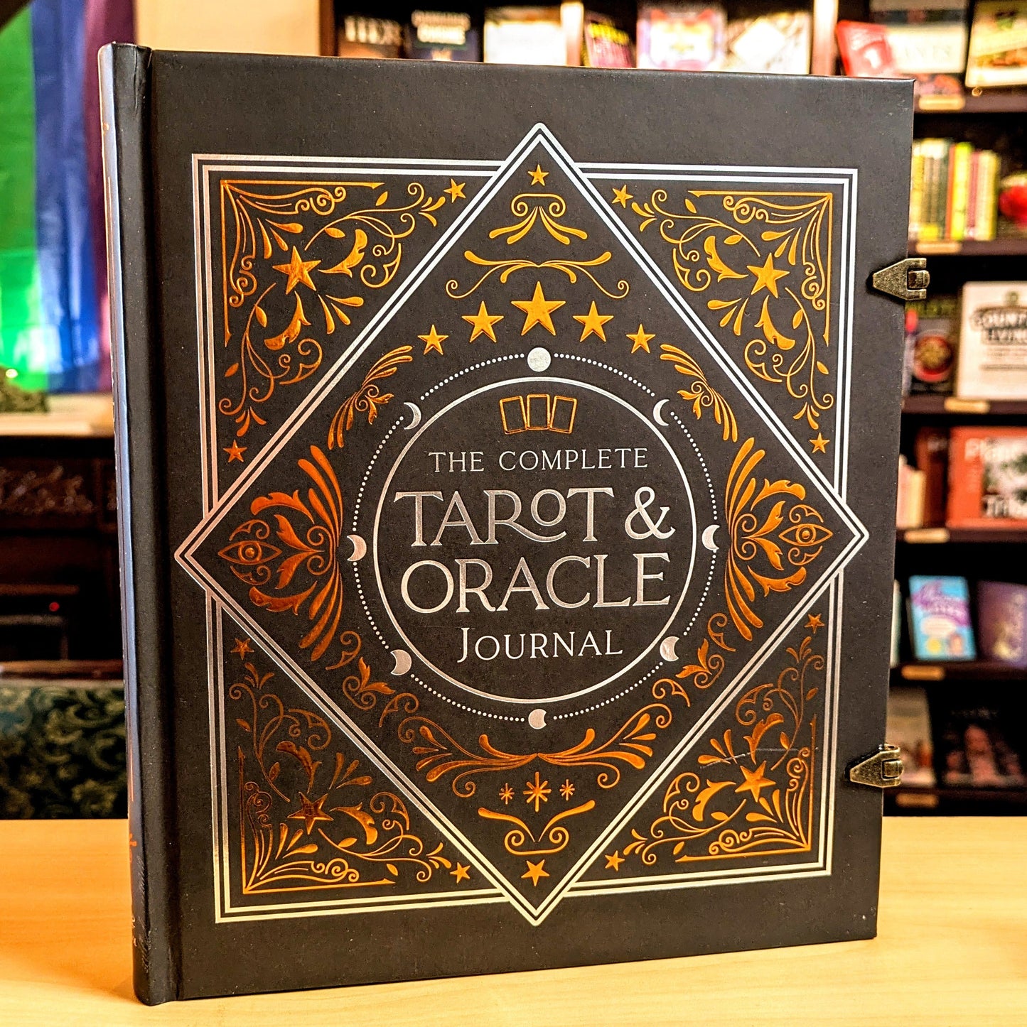The Complete Tarot & Oracle Journal standing upright on a wooden surface in a bookstore setting. The black hardcover features ornate gold and white decorative border designs with stars, moons, and filigree, and the title is clearly visible in the center.