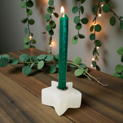 A lit green taper candle in a white star-shaped holder, placed on a wooden surface with eucalyptus branches and warm string lights in the background.
