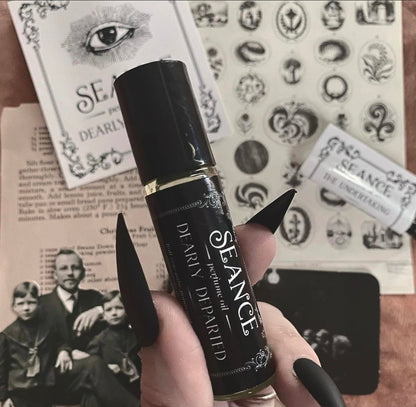 A hand with long black fingernails holds a black cylindrical perfume rollerball bottle labeled 'SEANCE' and 'Soul of the Spirit'. The background is a styled flat-lay of vintage-style papers featuring occult illustrations, tarot cards, and text.
