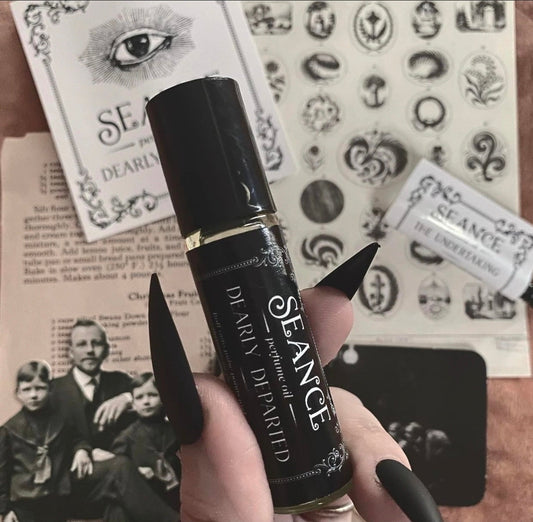 A hand with long black fingernails holds a black cylindrical perfume rollerball bottle labeled 'SEANCE' and 'Soul of the Spirit'. The background is a styled flat-lay of vintage-style papers featuring occult illustrations, tarot cards, and text.