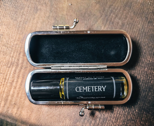 A perfume oil bottle labeled 'CEMETERY' resting inside an open silver-toned metal case with a black velvet interior, placed on a brown wooden surface.