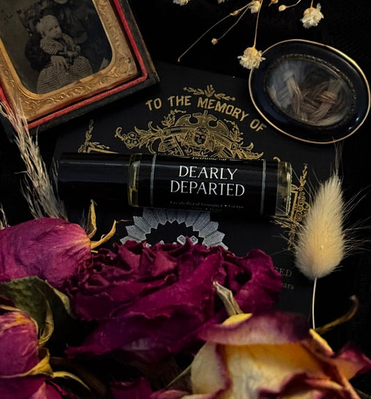 A black cosmetic tube labeled 'DEARLY DEPARTED' with gold lettering, styled on a dark surface with dried flowers, a vintage frame, and a round mirror.