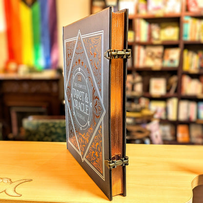 The Complete Tarot & Oracle Card Deck standing upright on a wooden table, with its ornate dark gray cover and brass-colored metal hinges visible, set against a blurred bookshelf background.