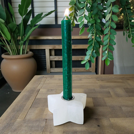 A lit dark green pillar candle in a white star-shaped holder, set on a wooden table with potted plants in the background.