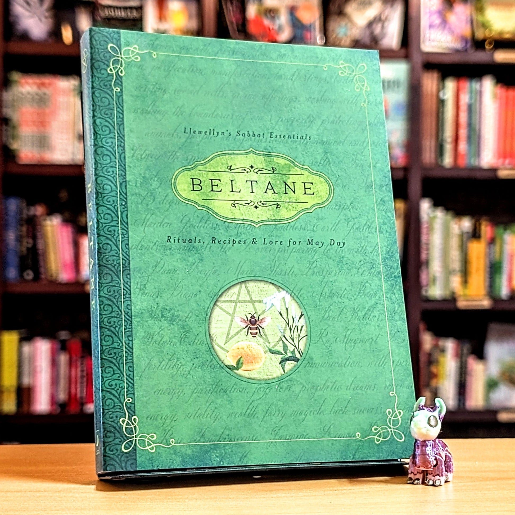 Green book titled 'Beltane' with decorative elements on a wooden surface in front of a bookshelf.