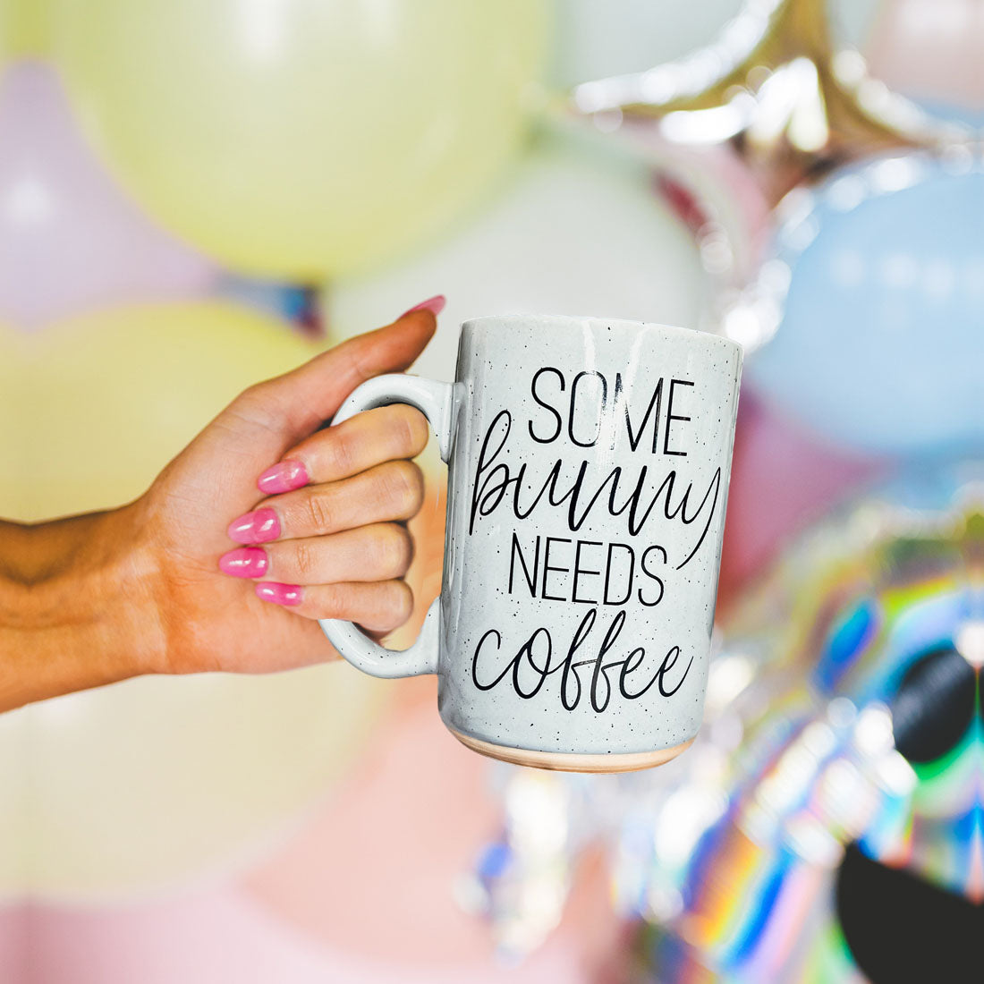 Hand holding a mug with 'Some bunny needs coffee' text against a colorful balloon background