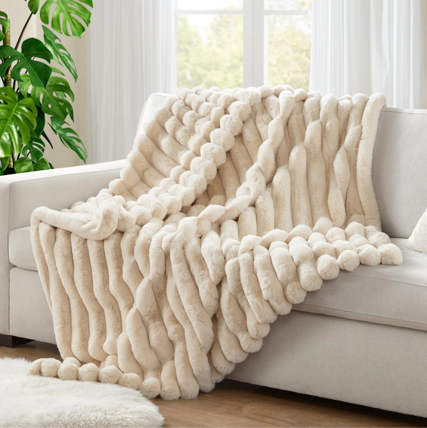 Beige textured blanket draped over a sofa in a bright living room with a plant and window in the background.