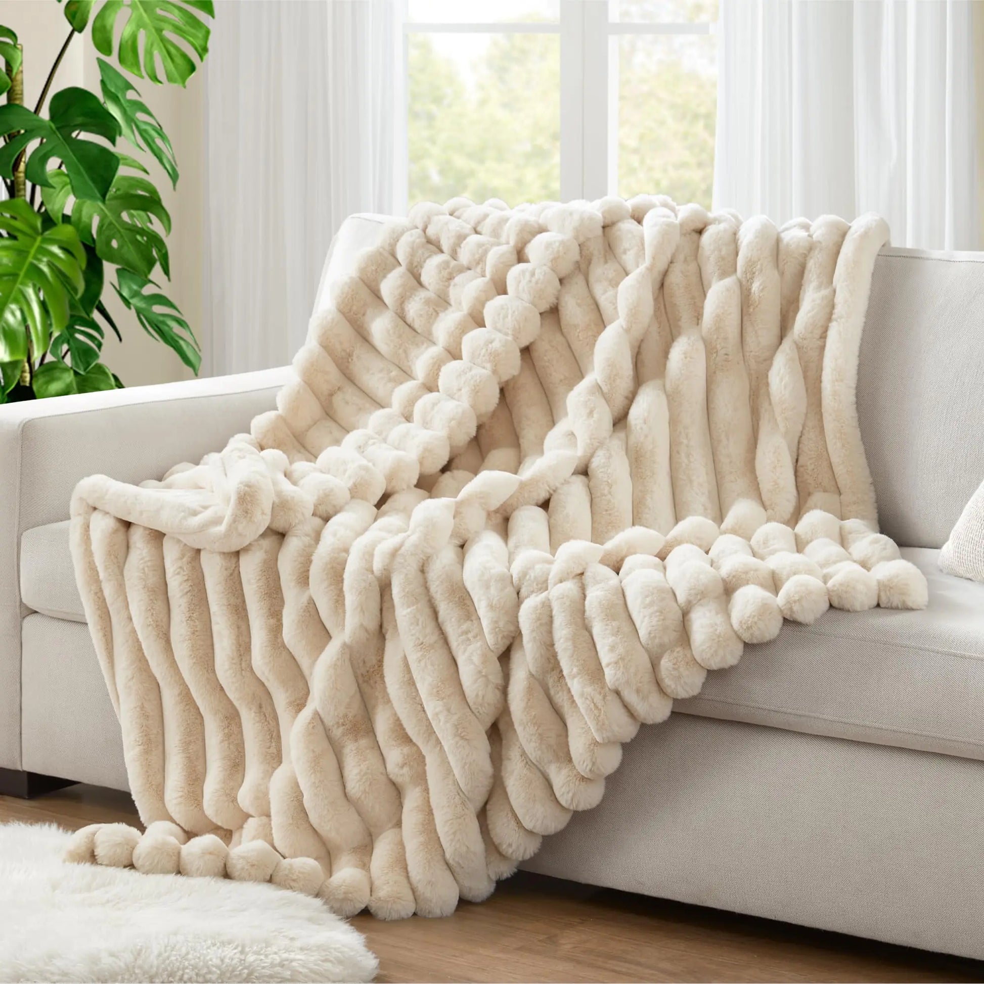 Beige textured blanket draped over a sofa in a bright living room with a plant and window in the background.
