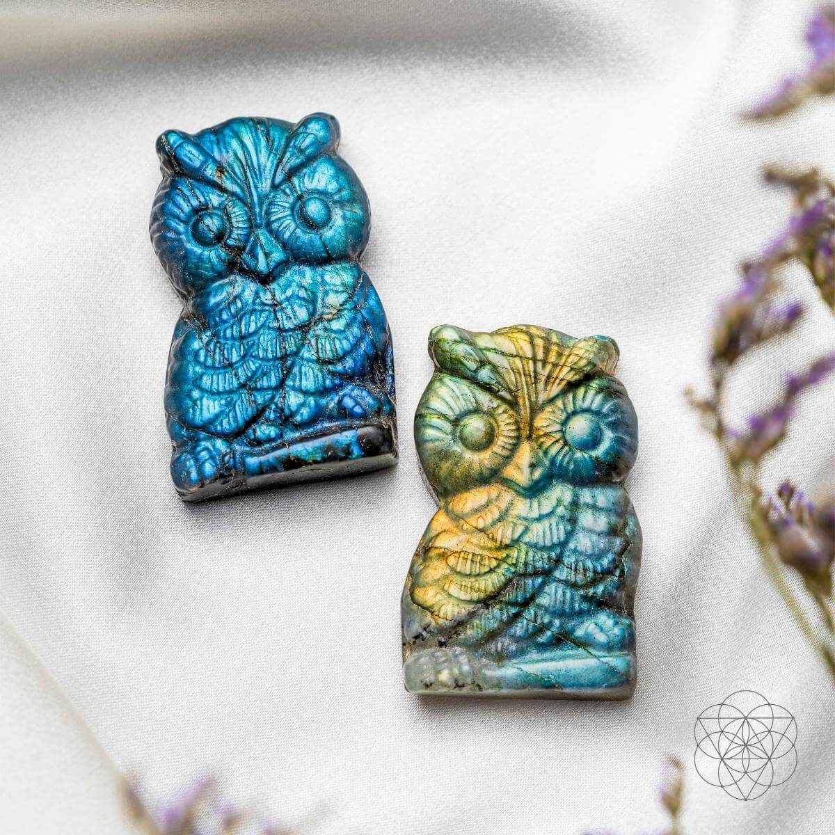 Two ceramic owl figurines with blue and green glaze on a white surface with purple flowers.