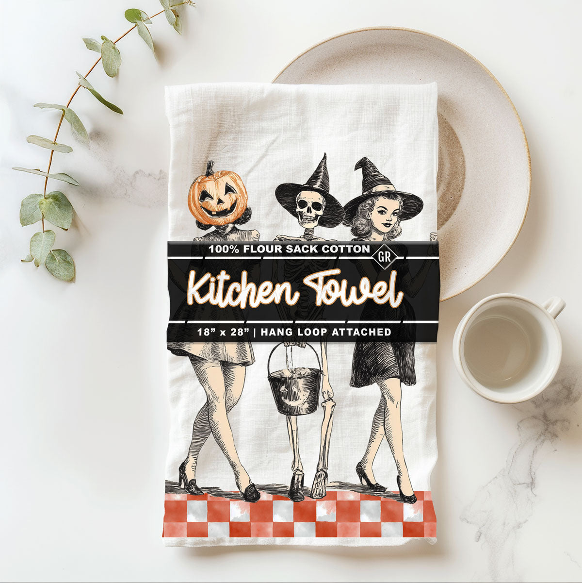 Halloween-themed kitchen towel with illustrations of a witch, pumpkin, and skeleton on a white marble surface.
