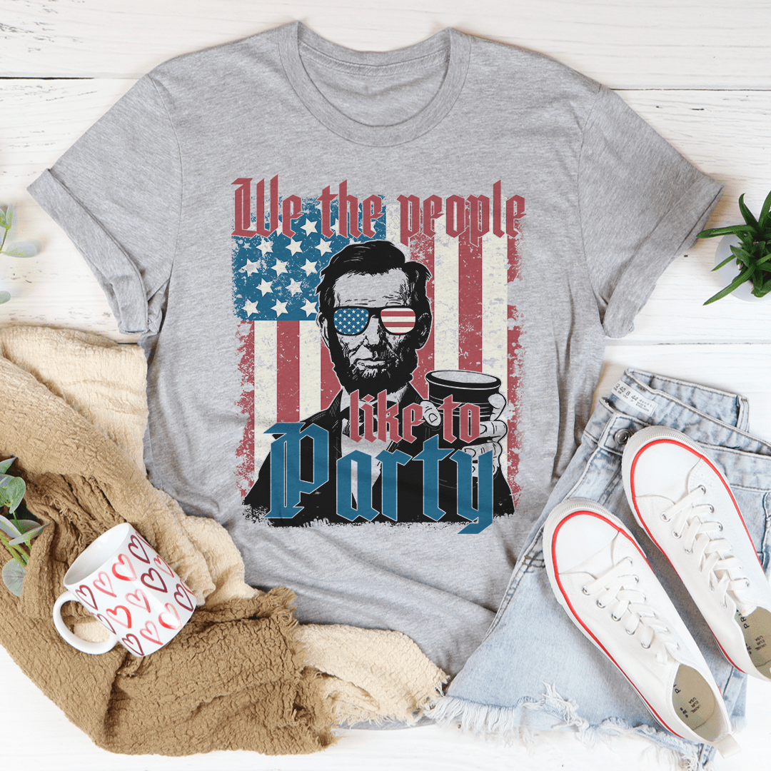 Gray t-shirt with a graphic of Abraham Lincoln holding a coffee cup and text 'We the people like to party' on a wooden surface.