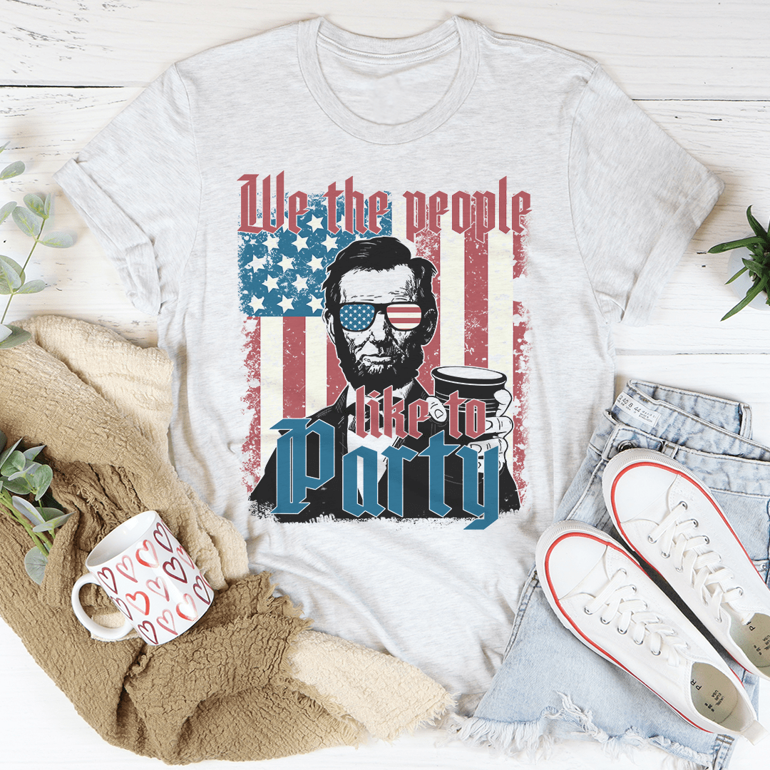 T-shirt with a graphic of a historical figure wearing sunglasses and an American flag, surrounded by casual items on a wooden surface.