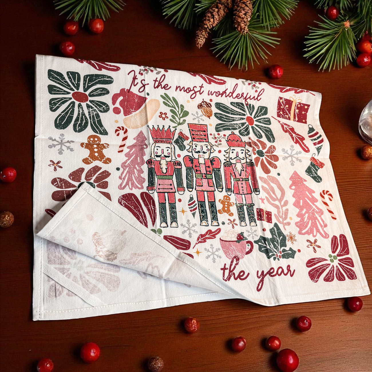 Christmas-themed kitchen towel with nutcrackers and festive text on a wooden surface with decorations.