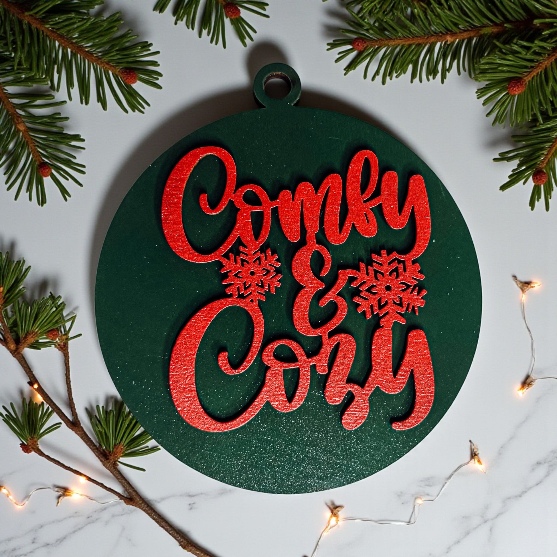 Decorative ornament with 'Comfy & Cozy' text surrounded by Christmas decorations on a marble surface.