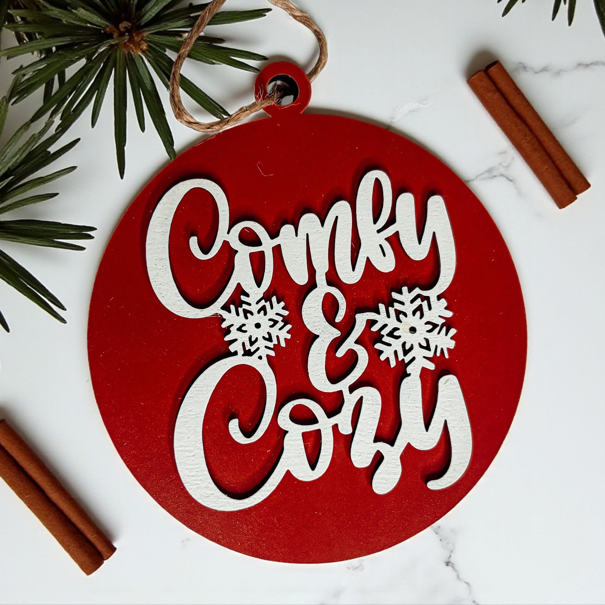 Red circular ornament with 'Comfy & Cozy' text and snowflakes on a white background with cinnamon sticks and pine branches.