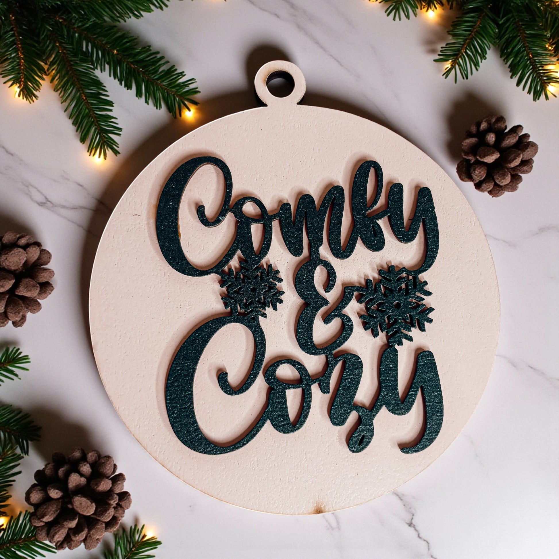 Decorative wooden ornament with 'Comfy & Cozy' text surrounded by Christmas decorations on a marble surface.
