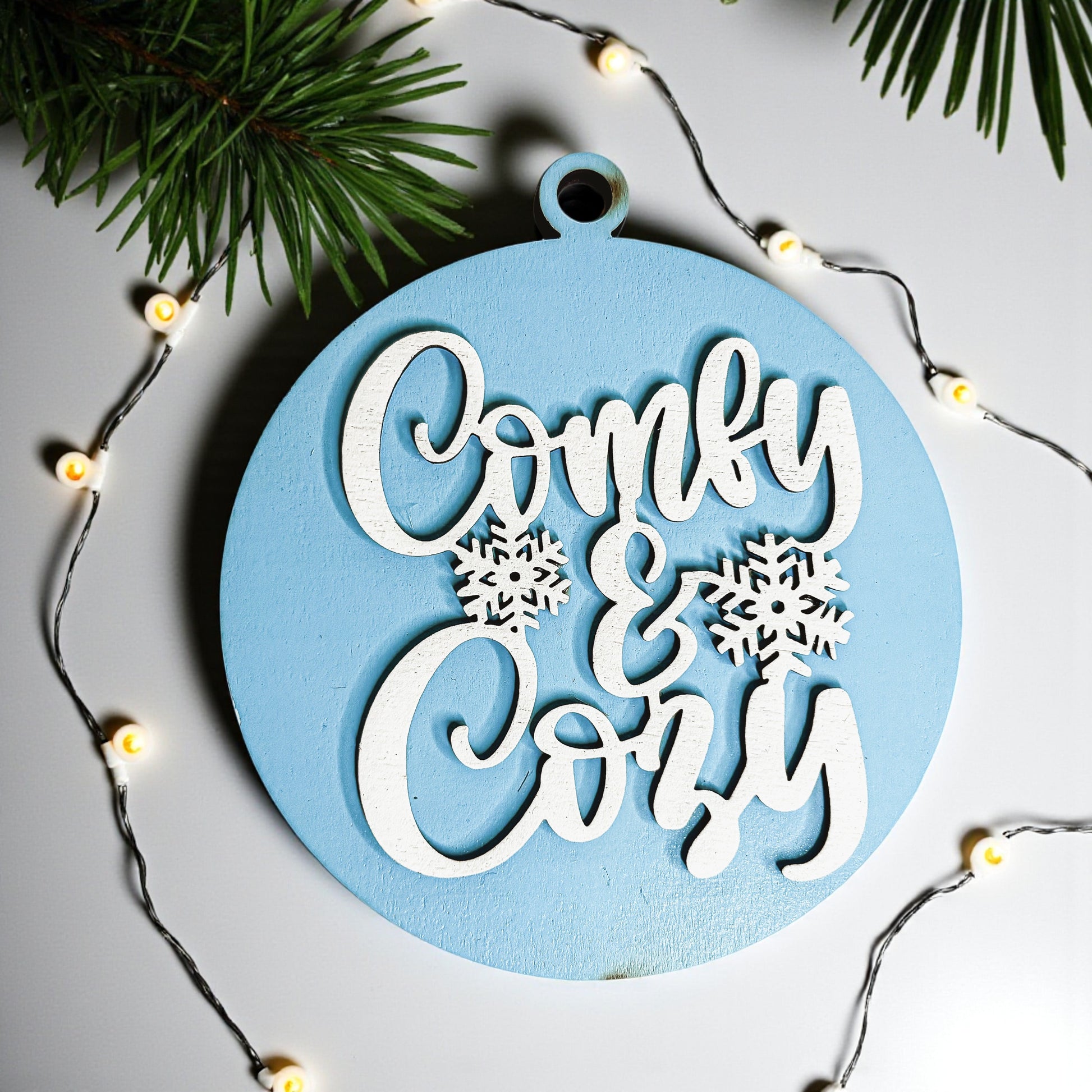 Decorative ornament with 'Comfy & Cozy' text and snowflakes on a light blue background, surrounded by Christmas lights and greenery.