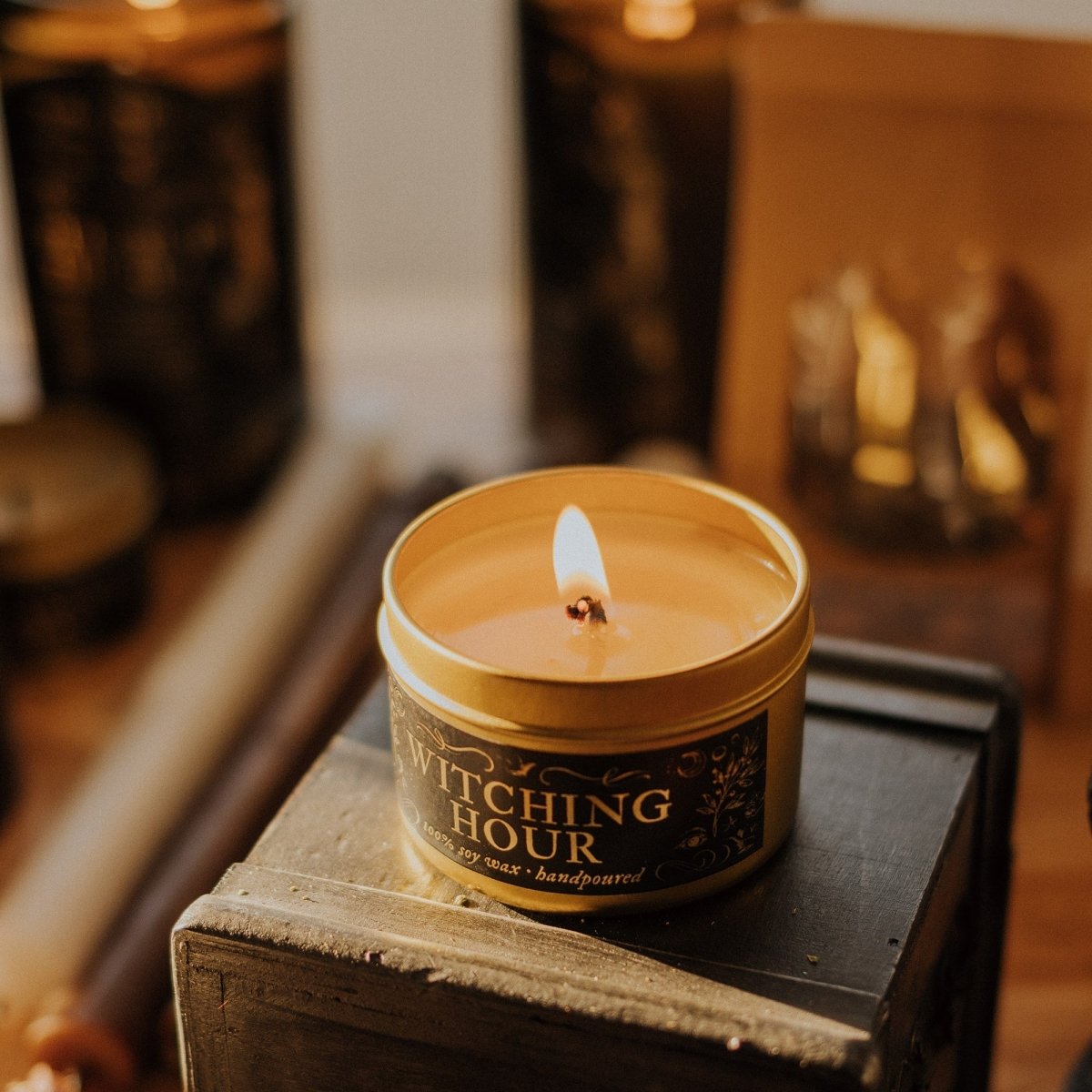 Candle in a tin labeled 'Witching Hour' on a wooden surface with a warm glow.