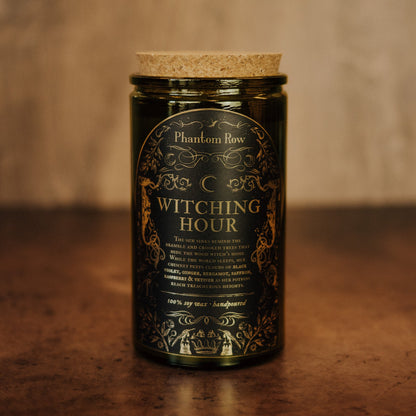 A dark green glass jar candle with a cork lid and a gold ornate label reading 'Phantom Row' and 'Witching Hour,' set on a wooden surface against a warm blurred background.