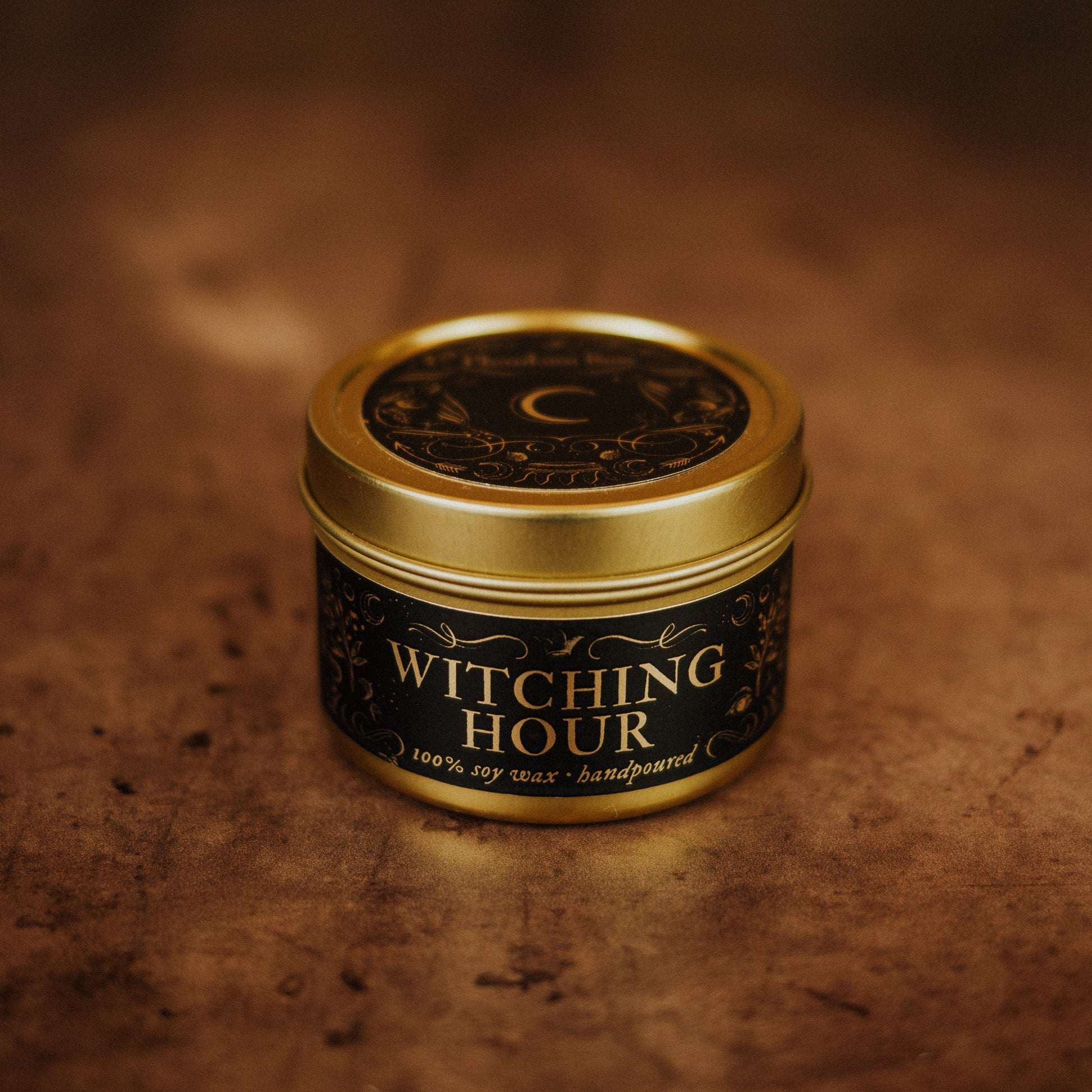 Candle in a decorative tin labeled 'Witching Hour' on a textured brown background